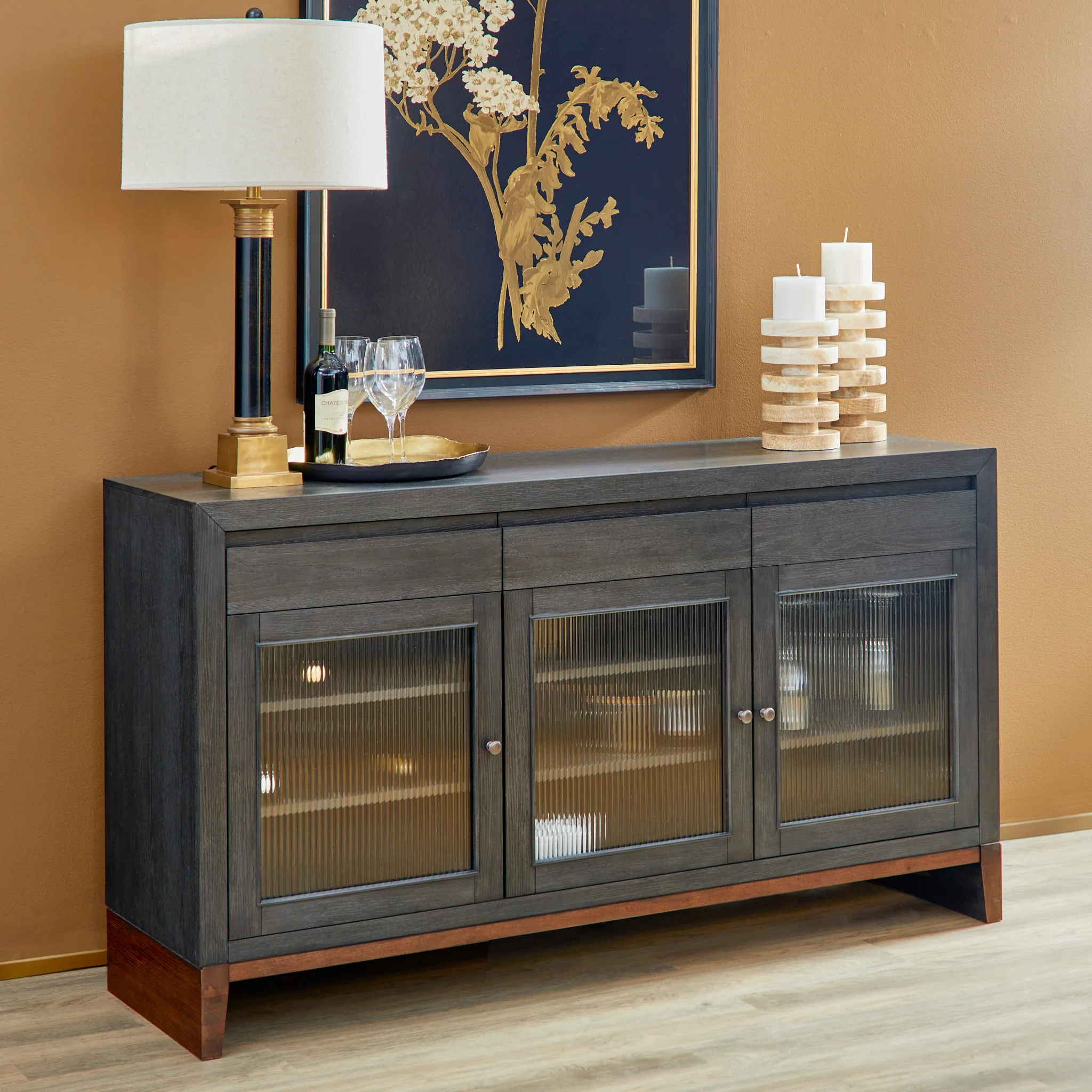 Flexsteel Waterfall Buffet in deep ebony and dark brown, measuring 65"W x 18"D x 36"H. Features three reeded glass front cabinets, wine storage for six bottles, wine glass racks for nine glasses, and antique copper hardware, combining style and functionality in a transitional design.