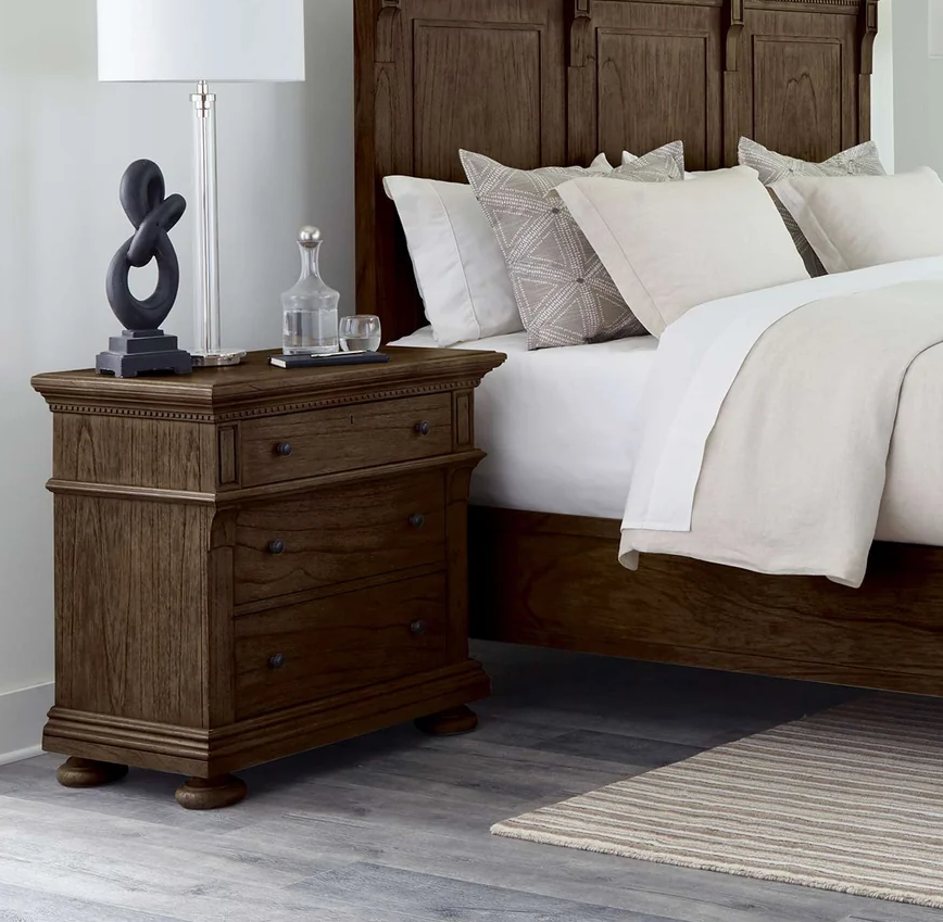 Hekman three-drawer nightstand crafted from Mindi veneers and select Mindi solids. Features self-closing slides, a cantilevered top drawer with corbel support, rustic antique brass hardware, and dentil molding in a Java finish.