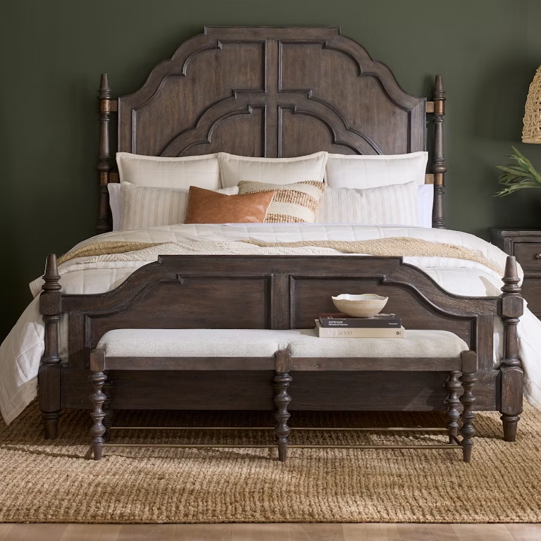 Rustic dark wood panel bed with intricately carved headboard, styled with neutral bedding, a textured rug, and a cozy bench at the foot.