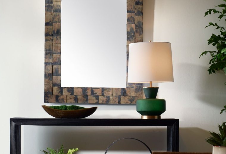 Rectangular mirror with a rustic wooden frame styled above a black console table with a green lamp, decor, and potted plants.