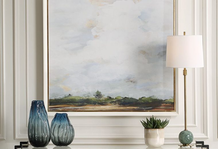 Serene landscape wall art with a gold frame styled above a console table with blue vases, a potted plant, and a gold lamp.